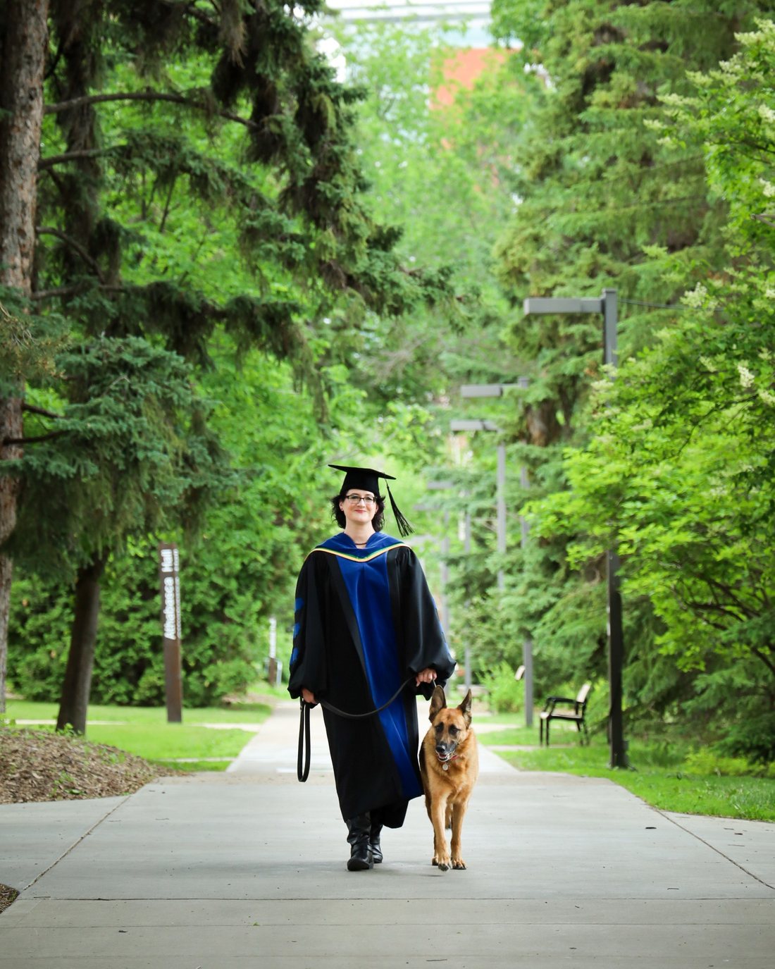 Sonja wearing academic regalia and walking with a dog on a tree-lined campus path.