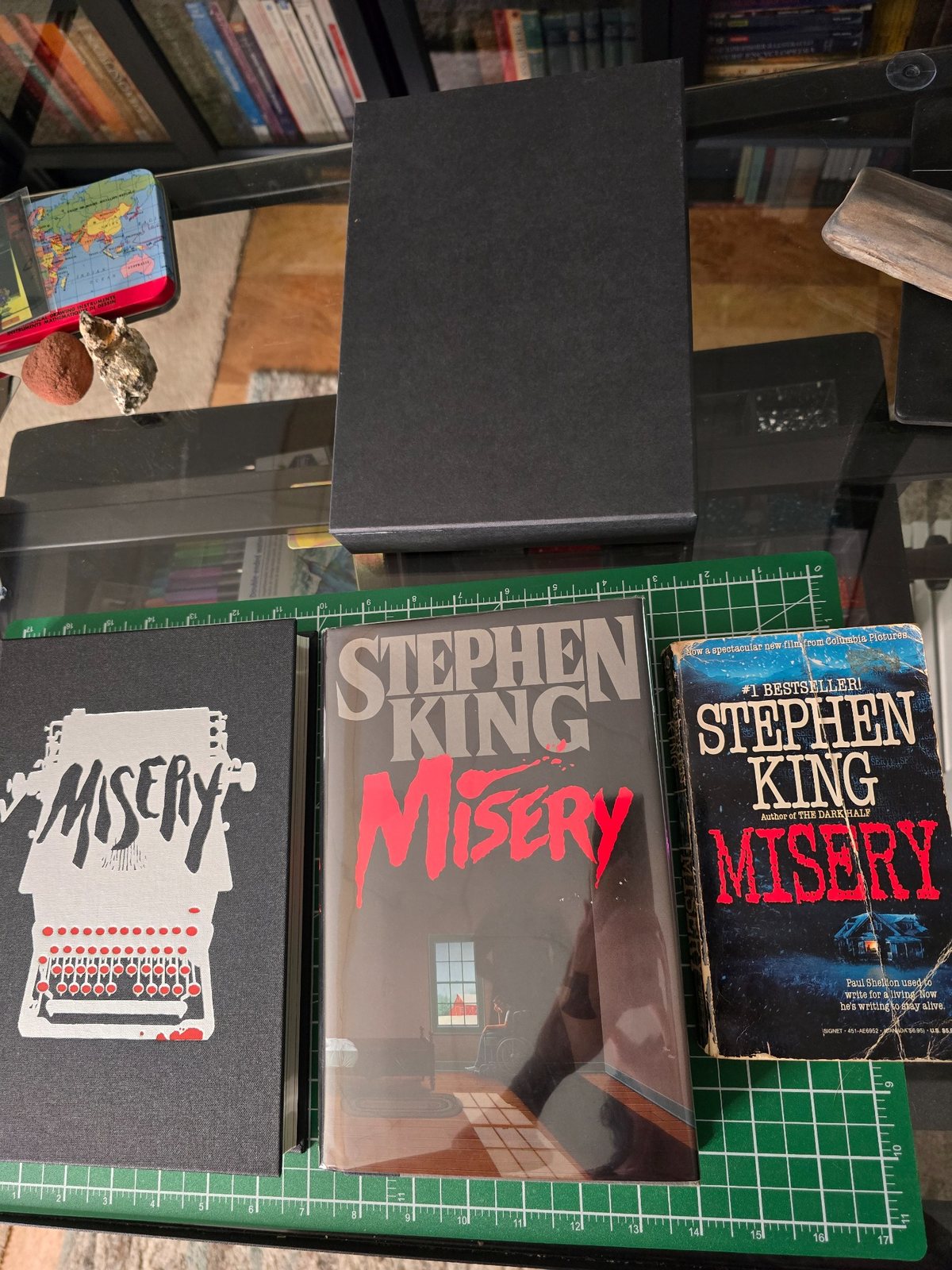 Several copies of Stephen King’s Misery arranged on a green cutting mat, including a signed first edition, a paperback, and a Folio Society edition.