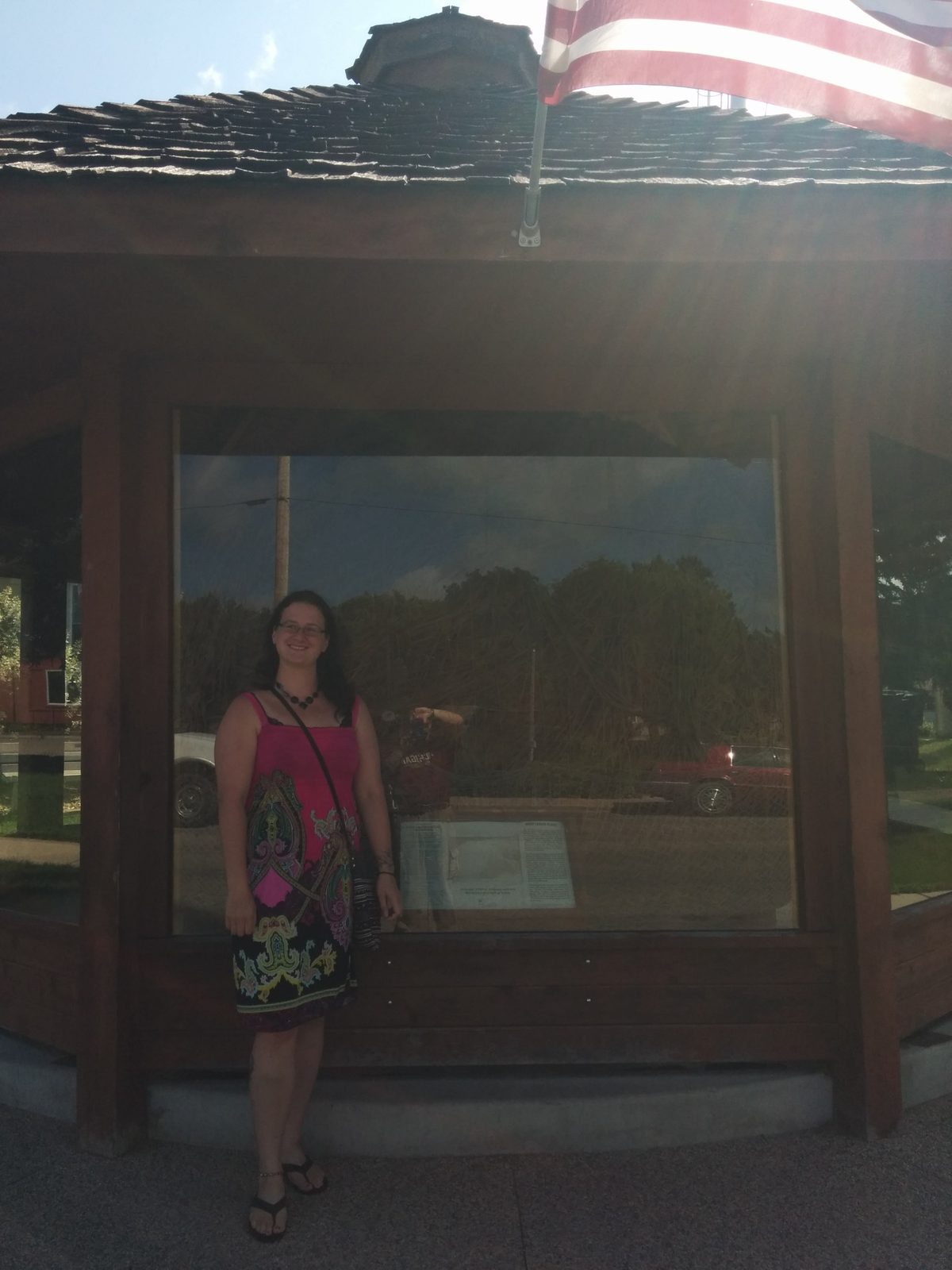 Sonja standing beside the glass enclosure containing the large ball of twine in Darwin, Minnesota, with an American flag above it.