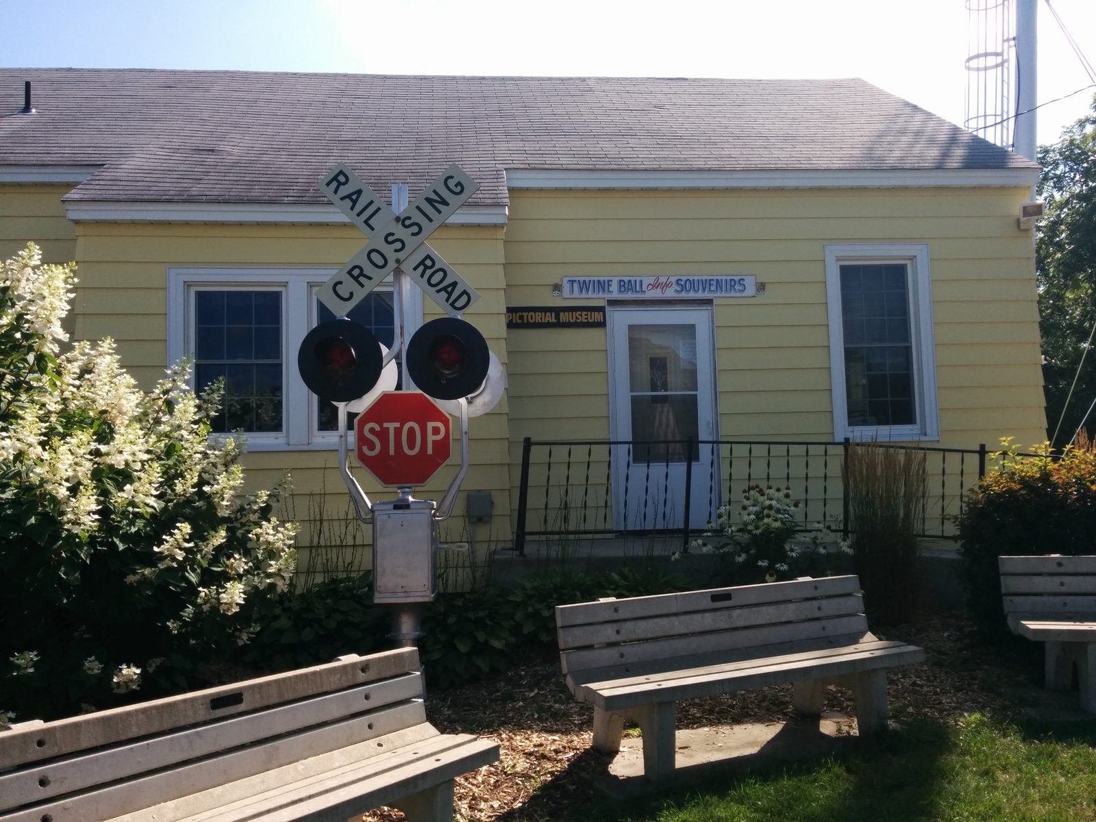 A yellow building with a sign reading Twine Ball Info Souvenirs, with a railroad crossing signal, stop sign, benches, and flowers in front.