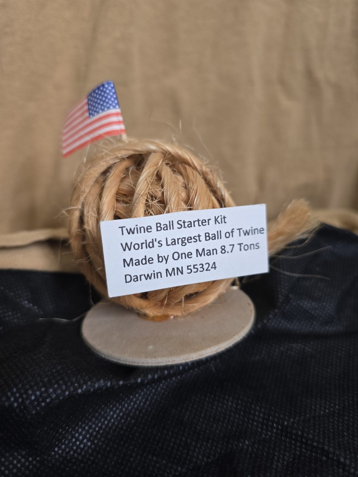 A miniature ball made from coarse brown twine sits on a round base with a small American flag and a white printed label identifying it as a Twine Ball Starter Kit from Darwin, Minnesota.