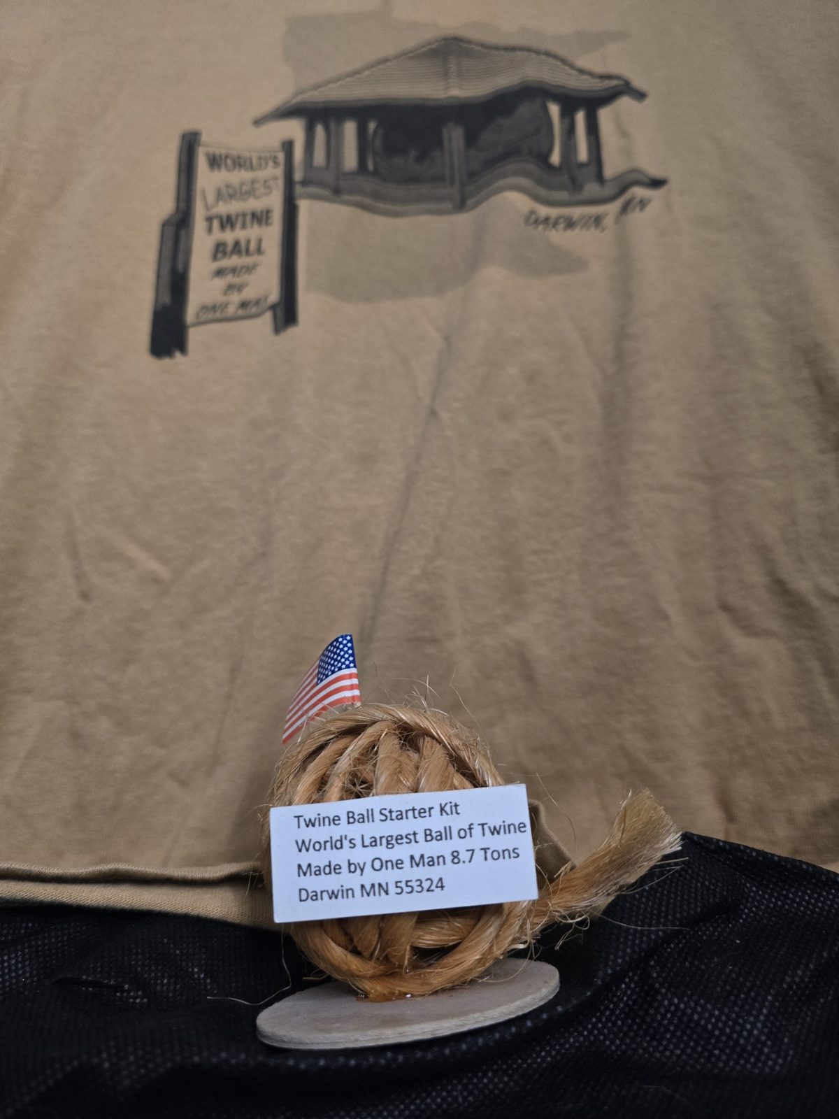 A small souvenir ball of twine with a miniature American flag and printed label sits in front of a tan T-shirt printed with the Darwin, Minnesota Twine Ball pavilion.