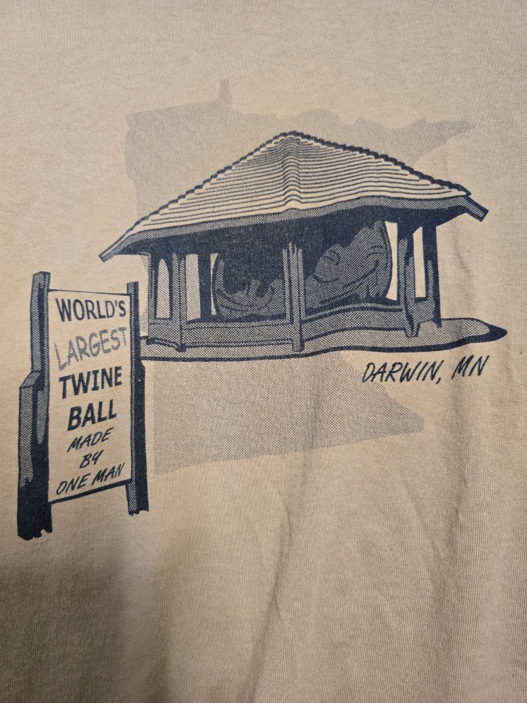 Close view of a tan T-shirt printed with an illustration of the Darwin, Minnesota Twine Ball pavilion and a sign reading World's Largest Twine Ball Made By One Man.